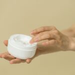 Close-up of female hands applying moisturizer from an open jar, highlighting skincare and self-care routine.